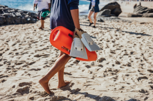 Man carrying a surfboard walks on the sand, symbolizing recovery and return to the water after heart surgery.